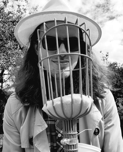 A person wearing a straw hat, sunglasses, and a trench coat, holding a fruit picker in front of their face, with an orchard in the background.