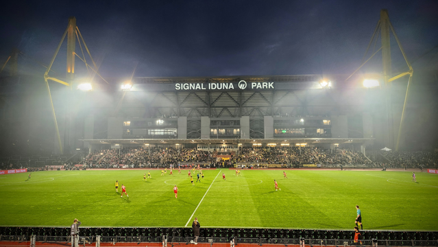Man sieht das Stadion Rote Erde in Dortmund und ein Flutlichtspiel. Um die Mittellinie herum sind die Spielerinnen beider Frauschaften im Spiel. Im Hintergrund die Osttribüne des Westfalenstadions.