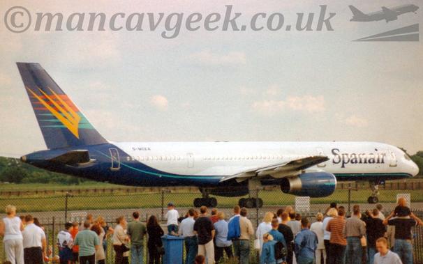 Side view of a twin engined jet airliner taxiing from left to right.
The plane is mostly white, with a dark blue belly, rear fuselage, and tail, with a green-blue pinstripe.
There are large blue, billboard-style "SpanAir" titles on the forward fuselage, and the black registration "G-MCEA" on the upper rear fuselage.
The tail has red, yellow, and greeny-blue spiky shapes, similar to forks, overlaid on each other.
The engine pods under the wings are also this dark blue.
In the foreground, a large crowd of people are standing in front of a tall, black, chain-link fence, mostly watching this plane.
Green grass is visible in the background under the plane, leading up to trees in the distance.
Very pale sky with fluffs of white cloud fills the rest of the frame.
