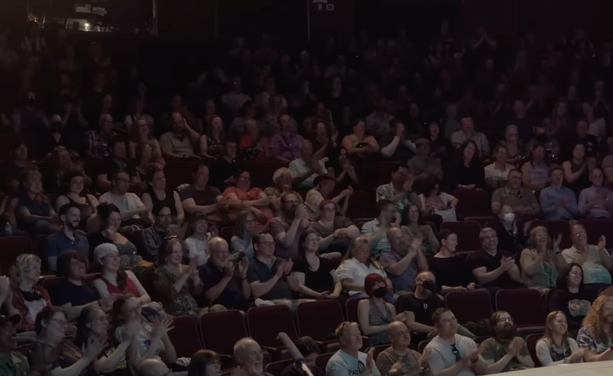 screenshot from the video linked in toot. We see a seated audience. There are three people wearing respirators. Two of those people are sitting in the second row, right in the center.