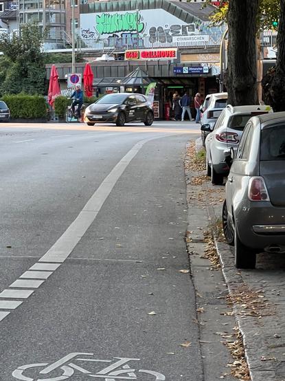 Foto des alten Radstreifens in der Reventlowstraße im Bereich kurz vor der S-Bahn-Brücke. Der Streifen ist recht schmal rechts daneben parken Autos, es gibt keinen markierten Sicherheitstrennstreifen.