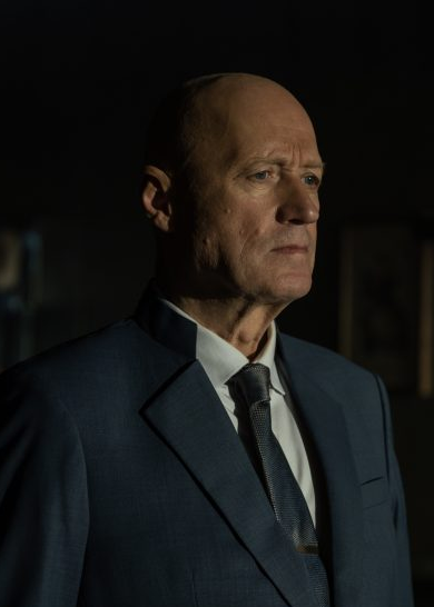 Image of character Atom Eins played by Adrian Edmondson from the TV series "Alien Earth".
A stern-looking bald white man with a heroic nose is show in partial light. He is wearing a dark suit, white shirt and dark tie.