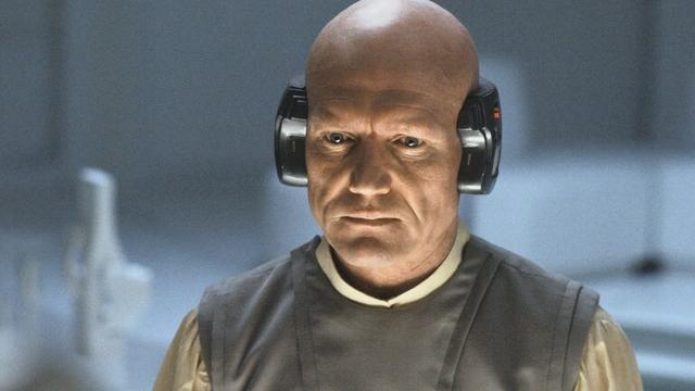 Image of character Lobot played by John Hollis from the movie "Star Wars: The Empire Strikes Back".
A stern-looking bald white man with a heroic nose is show in partial light. He is wearing a dark tunic over a pale shirt and has electronic devices attached to his ears..