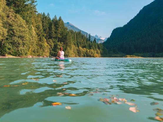 A woman in a bikini sits on a SUP in the middle of a glacial lake surrounded by fir trees on a sunny warm autumn day.