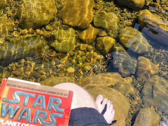 Someone reading a book with their feet in a rock-filled lake. The book is a Star Wars graphic novel.
