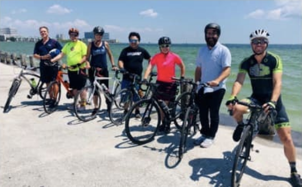 Photo credit:  Love to ride.  A group of people standing on a pier in front of the sea, with bikes.  They are wearing cycle clothing and helmets.