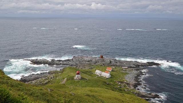 A photo looking down a green hill at a small lighthouse and several other buildings on a jagged rocky coastline. The sky is cloudy.