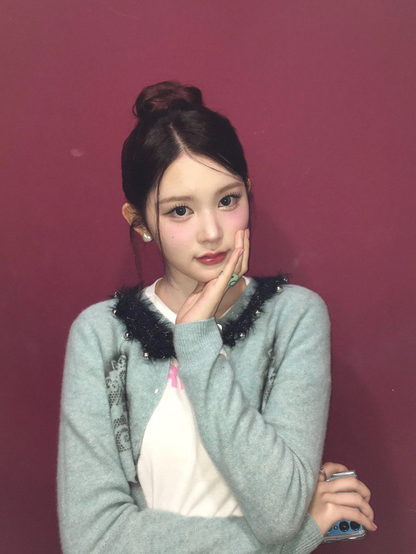 The image features a person standing against a solid maroon background. The individual is wearing a celadon cardigan with a black, furry collar adorned with small silver beads. Underneath, a white t-shirt with a pink design is visible. The person has their hair styled in a bun and is wearing pearl earrings. They are holding a smartphone with a blue case in their right hand, and their left hand is resting on their chin. The overall composition is simple, with the person centered in the frame, drawing attention to their attire and accessories.

Provided by @altbot,  edited by @jemmesedi.