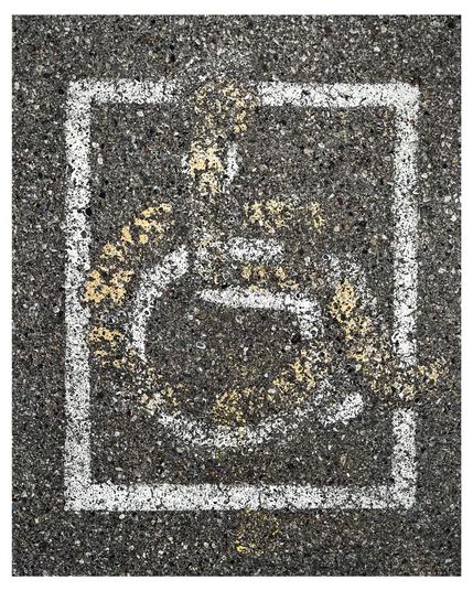 Faded white painted disabled parking stencil enclosed in a square on a textured asphalt surface, showing signs of wear and partially covered by a faded yellow stencil of slightly different design.