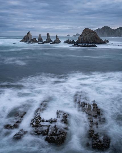 Rocky coastline with jagged formations emerging from the sea. Waves create a misty effect around the rocks, with overcast skies enhancing the moody atmosphere. Distant cliffs and scattered rocks further in the background.