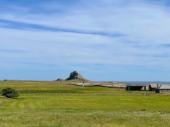 Lindisfarne, also known as Holy Island near Berwick-upon-Tweed in England, features open green fields leading toward the prominent Lindisfarne Castle, perched atop a rocky hill overlooking the North Sea. The sky is mostly clear with soft streaks of clouds, and the grassy landscape is dotted with a few stone buildings and fences in the foreground. A small group of visitors can be seen walking along the path toward the castle, emphasizing the island’s quiet and remote setting. The surrounding scenery combines coastal and rural elements, with the distant horizon meeting the calm sea.