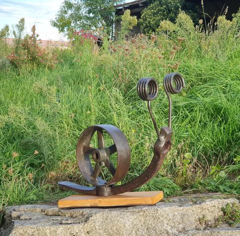 Sculpture escargot composée de pioches pour le pied, vieille roue métallique pour la coquille, ressorts de herse agricole pour les yeux, sur fond d'herbes vertes (socle planchette en pin)