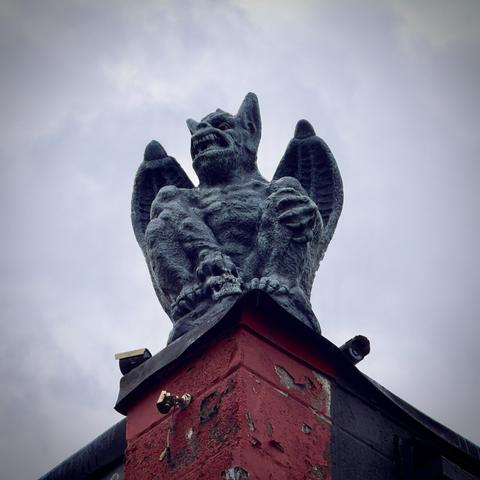 A deep gray gargoyle stands on the corner of a red and black concrete building, a skull clutched in one of its hands, under an overcast sky.