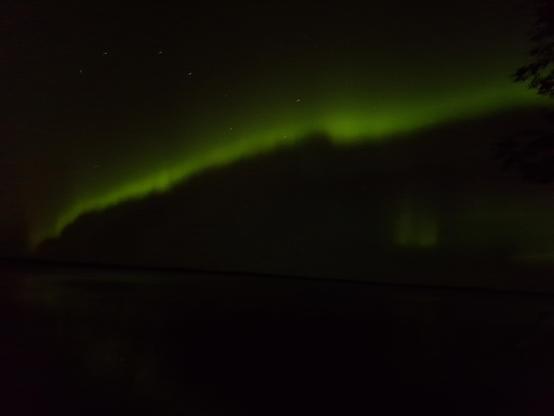 Image is a bit unfocused, but depicts very bright green Northern Lights dancing across the black sky above the lake.