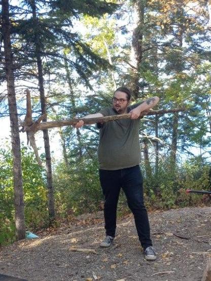 Image depicts Taylor, my spouse, using his found 'Gandalf Staff' with his battle face (which isn't very scary), and the background is trees and beyond that, the lake. He is wearing a green shirt and black skinny jeans, black Converse Chuck-style shoes, black and blue glasses, facial hair, snake bite lip piercings, and his hair is pulled back in a bun/pony.