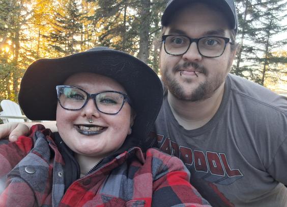 Image depicts me (left) wearing a black and red plaid jacket, a black witch hat, blue and black cat eye glasses, facial piercings, and black lipstick, and my spouse, Taylor, (right) with facial hair, snake bite lip piercings, black and blue glasses, a black ball cap, and a gray DeadPool shirt. We are smiling, and he has his arm around me. You can see trees in the background.