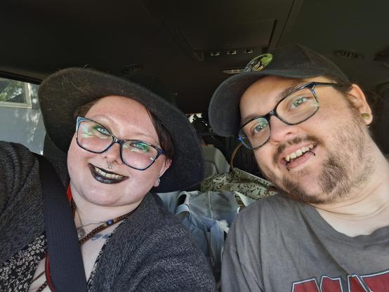 Image depicts me (left) wearing a black witch hat, black and blue cat eye glasses, black lipstick and eye makeup, a gray sweater, and a black and floral shirt, and my spouse, Taylor, (right) wearing a gray DeadPool shirt, black and blue glasses, snake bite lip piercings, and a black ball cap. We are leaning towards each other and smiling with our seatbelts on.