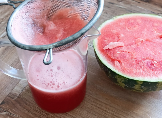 Ein Krug mit Melonensaft, durch ein Sieb gegossen, daneben eine halbe Wassermelone