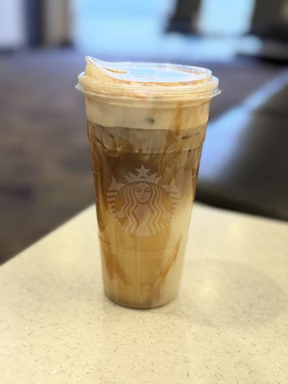 The image features a clear plastic cup with a lid, prominently displaying the Starbucks logo on the front. The cup contains a layered iced coffee beverage, with a distinct separation between the darker coffee at the bottom and the lighter milk or cream at the top. The layers are slightly blurred, suggesting a creamy texture. The cup is placed on a light-colored, speckled countertop, with a blurred background that hints at an indoor setting, possibly a café. The lid of the cup is slightly ajar, with a small amount of liquid visible on the rim, indicating the drink is ready to be enjoyed.