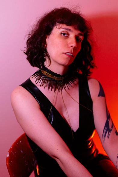 A person with curly dark hair wears a black sleeveless outfit and a spiked necklace, sitting on a chair against a pink and orange lit background.