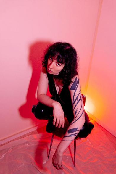 Un portrait serré, baigné d’une lumière rose et rouge. La personne porte une tenue noire brillante et un collier imposant aux pointes dorées. Son regard direct, légèrement de biais, et son expression calme ajoutent une intensité magnétique à l’image.