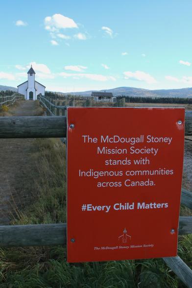 Photo of a sign on a fence post reading: The McDougall Stoney Mission Society stands with Indigenous communities across Canada. #EveryChildMatters."

McDougall Memorial United Church can be seen  in the photo's background.