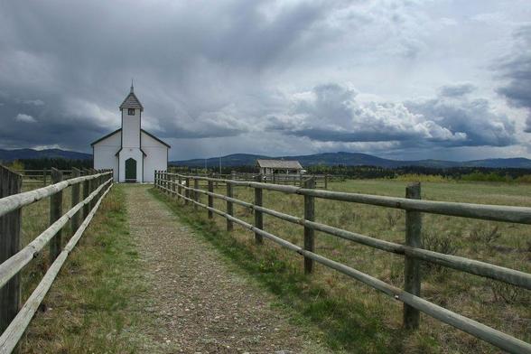 A small, very stereotypical-looking, white church stands at the end of a gravel lane, surrounded by green field. Simple wooden fences line each side of the lane, leading into the image and towards the church. Southern Alberta foothills spread across the horizon behind, and thick cumulous-like clouds hover in the sky, intuiting an oncoming thunderstorm.