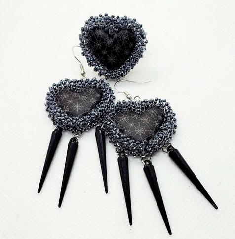 a pair of all-black heart-shaped beaded dangling earrings with a matching ring made by kiamichi. on the hearts are white thin cobweb designs which are surrounded by black shiny beads. from the bottom of the earrings hang 3 long black metal spikes