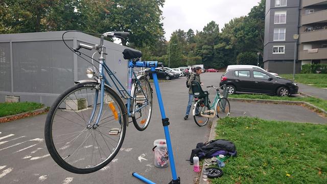 Sur un coin de parking dégagé de voitures par l'accès secours et un abri vélos :
- un chouette vélo MBK à col de cygne en fin de révision sur un pied d'atelier près d'un seau MucOff
- une femme dépose un vélo avec siège bébé.