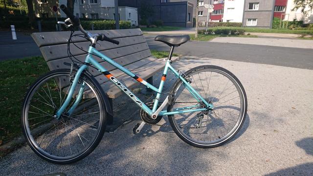 VTT mixte Orbéa émeraude monté pneus route au soleil après entretien, au pied d'immeubles.