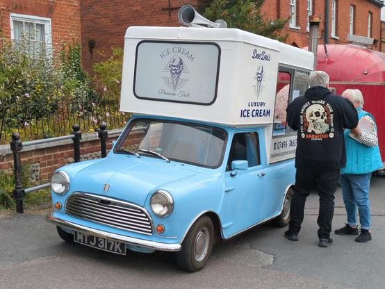 A tiny sky blue BMC Mini converted into an ice cream van. Signwritten 'Dee Dee's Luxury Ice Cream', it's a pick-up body (or a cut-down van) with a large white box on top which extends over the front of the cab and incorporates a serving window, in front of which an older couple are standing. Behind, a separate red Airstream-type caravan burger van can just be seen. They're parked at the edge of Reepham's market square, in front of some bollards and a low wall. Behind the wall, red-brick 18th century buildings provide an elegant backdrop.