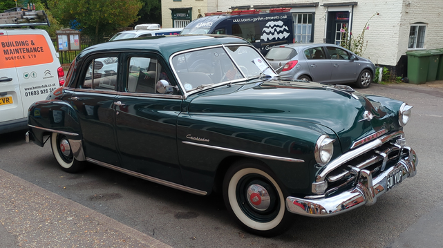 A beautiful curvy deep green Plymouth, dating from an era before extravagant fins. The front grille and bumper are large and chromed. The bumper has two huge chrome overriders and the grille appears to have three more. There are chrome strips along front and rear wings, chrome strips beneath all the windows, a chrome windscreen surround and a chrome bonnet strip. The hubcaps are - have a guess... yep - chrome, with a smaller circular red area, the better to see the chrome logos in the middle. Various modern vans and cars are in the background, in front of white-painted 18th century brick buildings. In the distance at the rear, a wooden notice board headed Reepham Town Council stands in front of a fence which surrounds an area of grass and trees.