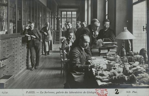 une grande salle avec sur la gauche un long mur comportant des tiroirs et des vitrines et à droite de la pièce des tables et des chaises avec des roches, des ouvrages et des hommes regardant le photographe