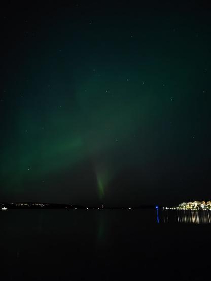 Turquoise northern lights circling above a lake. In the middle close to horizon there is more intense green northern lights with a hint of red color. Lake has a reflection of this intense line. On the right side of the horizon there is lots of lights from the city that the lake reflects as well. The sky is full of stars and on the right you can see the Big Dipper constellation.