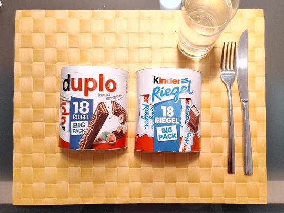 Foto: Eine Packung Ferrero Duplo und eine Packung Ferrero Kinder Riegel mit je 18 Riegeln auf einem Platzset, dazu Messer, Gabel und ein Glas Wasser.