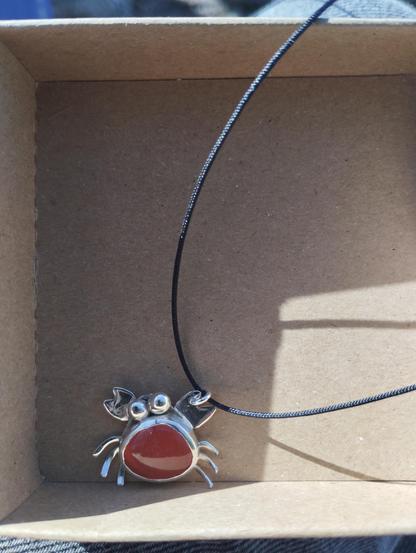 a silver pendant in shape of crab with an orange gemstone. it has a black nylon cord. it's in a carton box