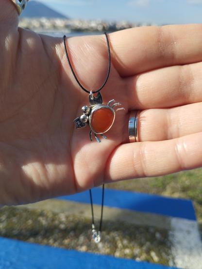 a silver pendant in shape of crab with an orange gemstone. it has a black nylon cord. it's hold by a hand