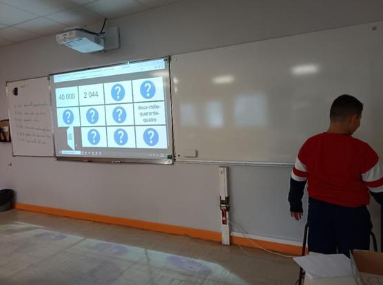 Elève devant un tableau, on voit un vidéo proche lecteur projeter un memory avec des nombres écrits en chiffres et en lettres.