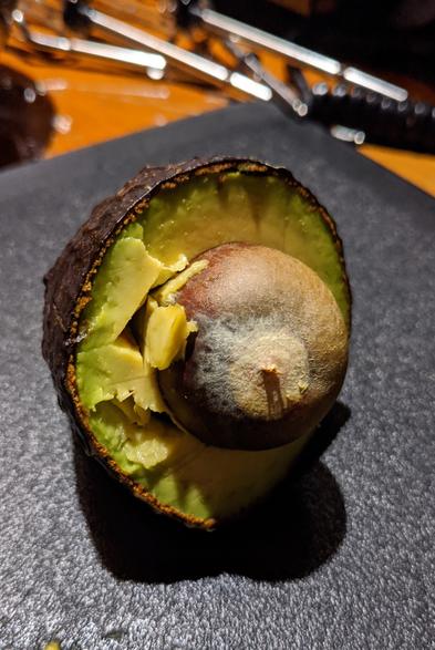 I hate avocados 🥑 

A halved avocado on a dark cutting board. The flesh is a vibrant green, with some darker spots, and the large, brown pit remains in the center. The skin is dark and textured. The background is out of focus, showing reflections of what appear to be kitchen tools or utensils.