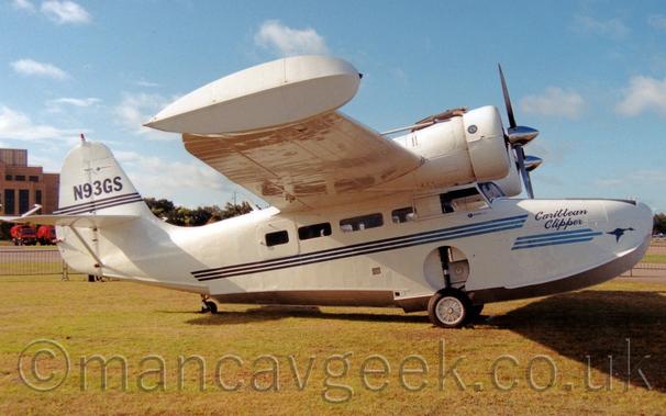 Side view of a white, twin propellor-engined amphibious plane parked on grass, facing to the right.
The plane is almost entirely white, with a trio of blue lines running along the body, with blue "Carribean Clipper" titles and an image of a flying goose on the nose.
The registration "N93GS" is on the top of the tail, above 3 more blue stripes.
The planes wheels , while deployed  for the plane to sit on, obviously retract into holes in the side of the fuselage, with the tail-wheel retracting into the tail boom.
The engines  sit forward of the high-mounted wings, close in to the fuselage, with retractable floats on the ends of the wings.
Green grass fills the foreground, with a brown brick building on the left of the background, under blue sky with wisps of white cloud.