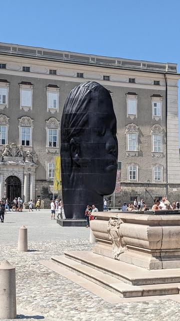 The stairs of a fountain on a public square are visible in the front. Many people of the public are visible around the fountain. In the distant a black head of a girl is visible. The shape is heavily scewed such that in this perspective the girl is recognizable, but the impression is that the shape is off due to the sun's light.
