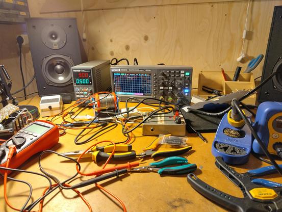 Picture of a desk with a lot of wires, tweezers and cutters, a multimeter, a soldering station, an oscilloscope and a dc power supply.