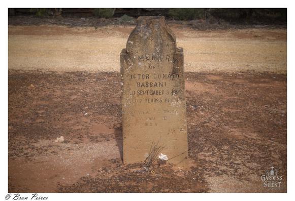 A close up photograph of a small, heavily weathered, rough-hewn sandstone headstone at the Kingston on Murray Cemetery.   The inscription is faded but mostly legible, reading: "MEMORY / OF / VICTOR DONATO / BASSANI / DIED SEPTEMBER 3 1897 / AGED 7 YEARS 5 MONTHS."  Below this, a faint epitaph begins "WE WERE NOT THERE..."  The ground surrounding the monument is composed of reddish-brown dirt and pale gravel, suggesting a dry, rural environment.
