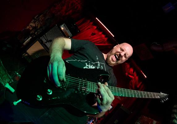 Gitarrist der Band "Raging Sloth" mit einem Fisheye-Objektiv fotografiert. Er steht auf der Bühne des Kunstvereins Nürnberg und zieht eine angestrengte Grimasse, während er Gitarre spielt.