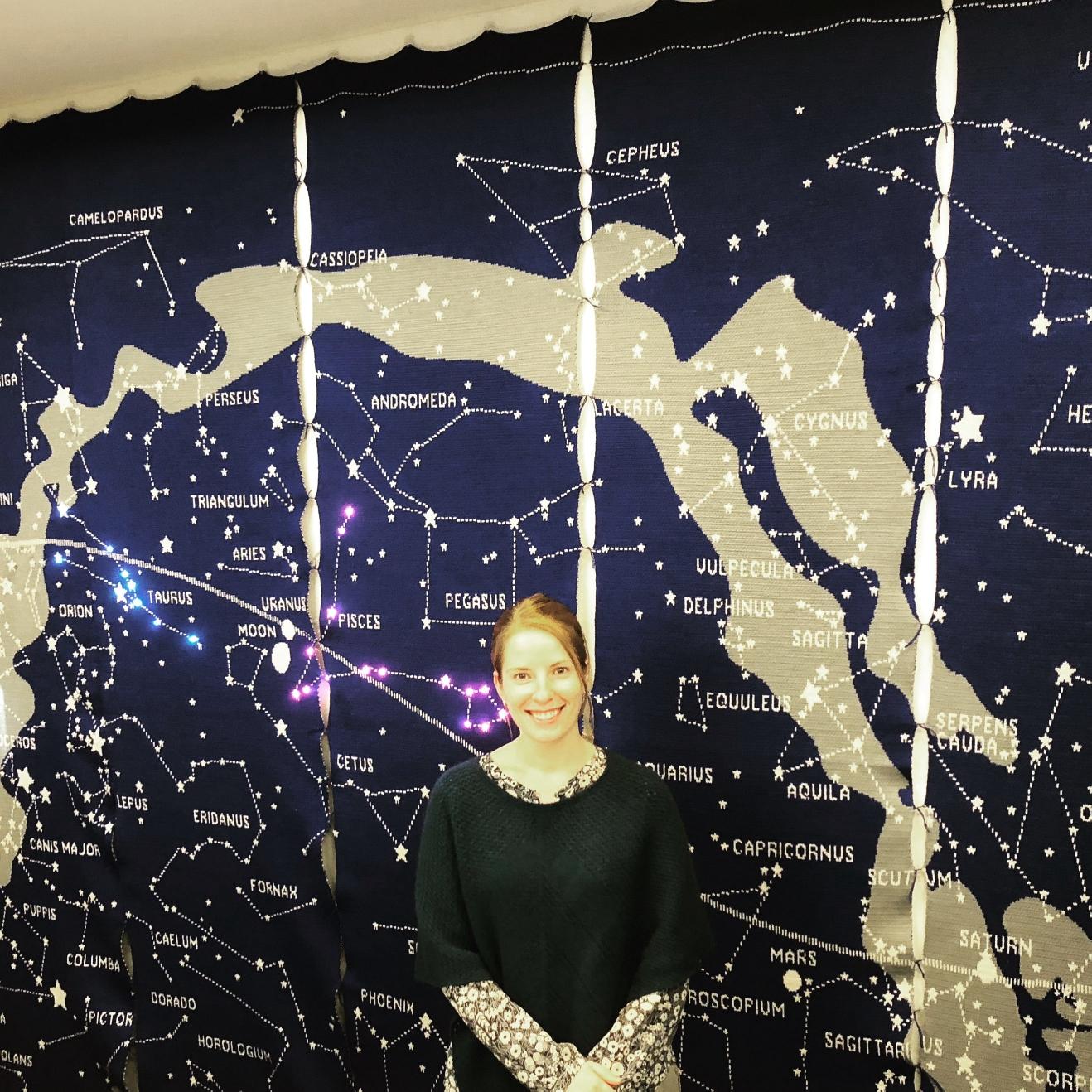 A woman standing in front of an enormous knitted map of the night sky, with constellations labelled