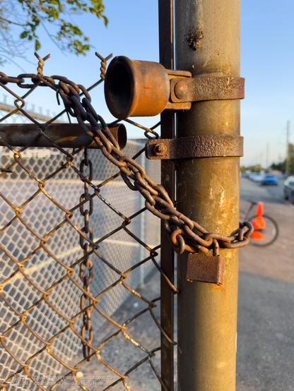 A rusty chain and padlock secure a chain-link fence to a metal post. An orange traffic cone and cars are visible in the background.