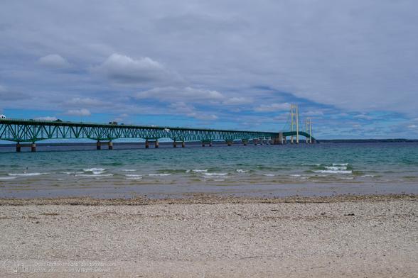A photograph of the Mackinac Bridge, a suspension bridge painted green, which spans the Straits of Mackinac, connecting Michigan's Upper and Lower Peninsulas. The bridge is viewed from a beach covered in small, light-colored stones and sand. Gentle waves lap at the shoreline. The water transitions from a clear, shallow area near the beach to a deeper, turquoise hue further out. Cars and trucks are visible traveling across the bridge. The sky is filled with a mix of blue and gray clouds, suggesting a partly cloudy day.