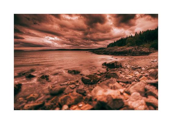 A monochromatic photo showing a rocky beach and forested headland looking out to sea with clouds low on the horizon. Shot with a red filter. Framed in white.