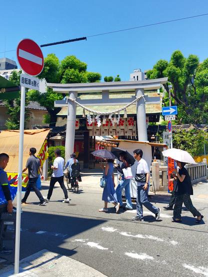 The main entrance to the shrine is located at the corner of an intersection. It was hot and I gave up in waiting to be able to get no passersby in the shot; it was busy that day. So I opted for most people in the shot instead. So there are about ten of them walking or standing, a couple holding parasols/umbrellas against the relentless sun. The gate itself is stone, a light gray in color, and it's decorated with a thick rope and paper streamers. It was matsuri/festival time at this shrine and the gate with doors behind the torii is adorned with red and white paper lanterns, as are the gazebo tents set up behind it lining the footpath to the shrine. The sky is perfectly blue.