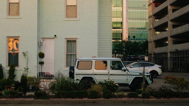 A street-level photo capturing a striking contrast in urban architecture. In the foreground, a Mercedes-Benz G-Wagon is parked in front of a brick two-story house, both painted a pale mint green. To the right of the house, the scene shifts to modern structures: a multi-story glass office building and a concrete parking garage. The warm, golden light of sunset is reflected in the windows of both the vehicle in the foreground and the house glass creating a moody and atmospheric scene.